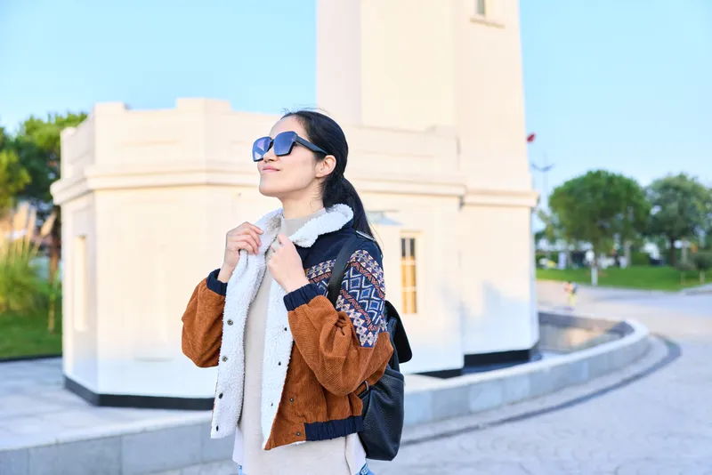 Woman wearing scarf and jacket with backpack, posing outdoors near a white monument, stylish street style shot highlighting fashion, travel vibe, and urban scenery