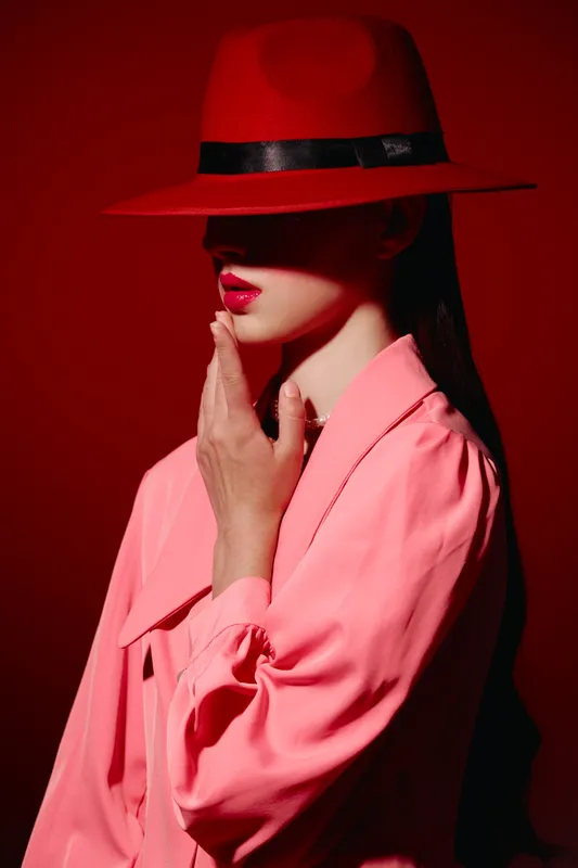 Fashion model in a red wide brim hat and pink jacket poses with lips painted bright red against a red background. Beauty and creative style concept with dramatic lighting.