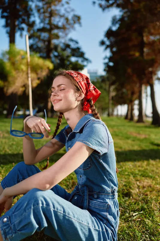 lifestyle woman in modern boho-western denim street style with red bandana sitting outdoors in natural sunny park with relaxed film color tones