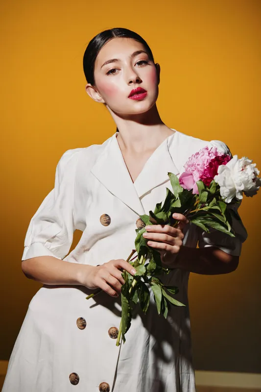 Young woman in white dress holding pink and white flowers poses confidently against mustard background. Bold red lipstick and sleek hairstyle highlight beauty and fashion theme.