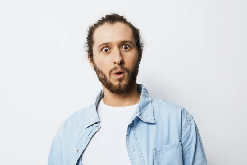 Surprised young man with a stylish beard and casual attire, showcasing an expression of shock against a minimalist light background