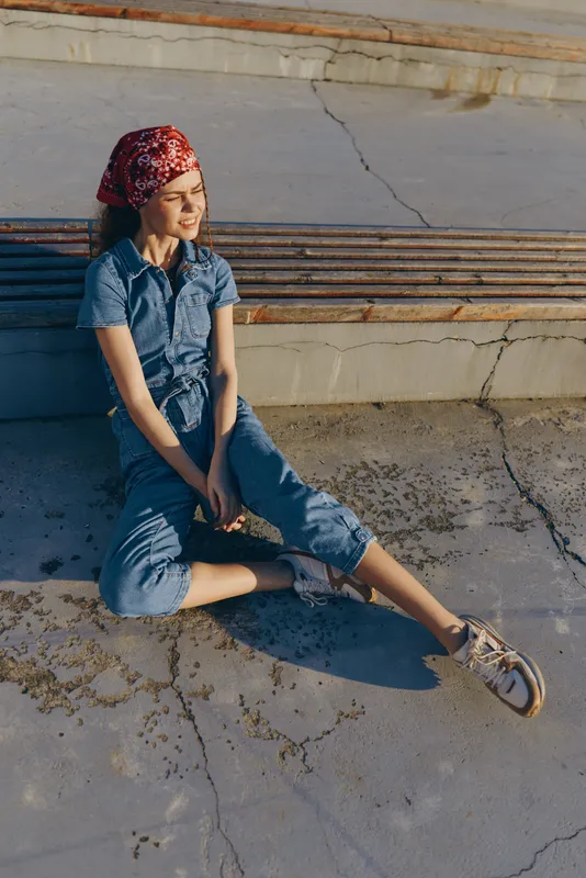 Young woman with red headscarf sitting on concrete floor in casual denim outfit enjoying sunlight and relaxed moment outdoors during daytime.