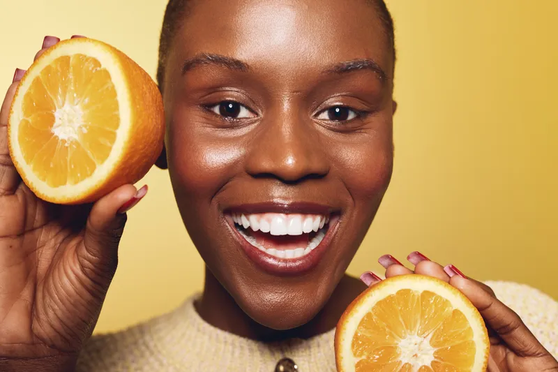 Happy African woman with dark skin holds two orange halves near face, smiling brightly on yellow background. Concept of healthy lifestyle, vitamin C, and natural beauty.