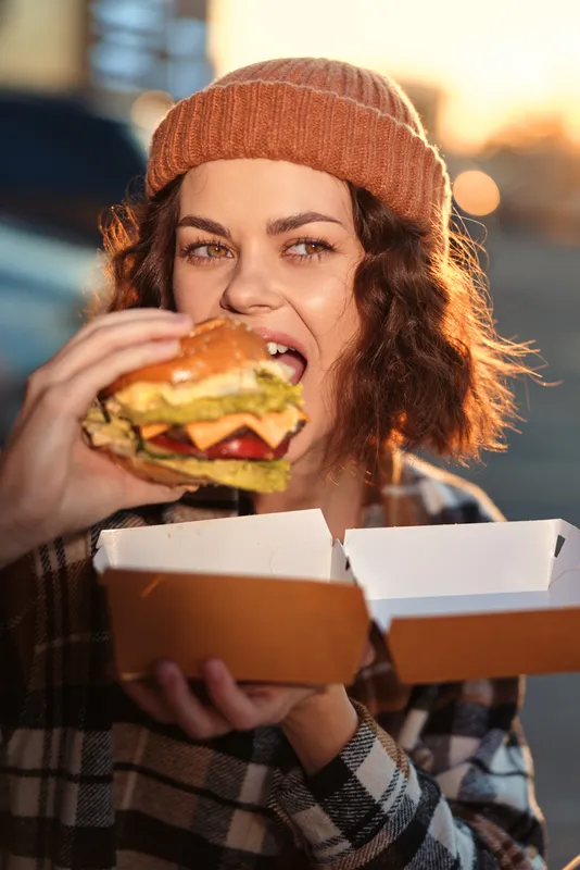 Woman eating a burger from takeout box during golden hour, candid urban portrait with beanie and authentic smile, emotional storytelling, mindful living and warm glow.