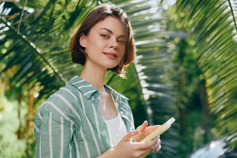woman outdoors smiles softly under sunlit leaves, wearing a striped shirt and white top, holding a phone, radiating confidence, curiosity, and a fresh natural mood for lifestyle and travel imagery