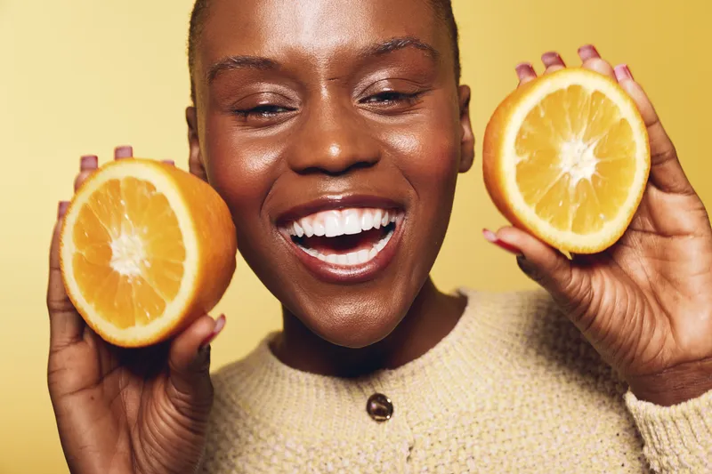 Smiling dark skinned African woman holds two halves of a ripe orange close to her face. Bright yellow background highlights fresh fruit and joyful natural beauty concept for health and wellness.