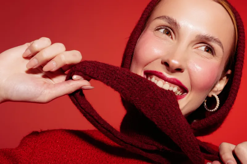 Smiling woman in red sweater and hood holds hood strings, looking away with bright lipstick and hoop earrings. Close-up on red background. Concept of fashion, beauty, and winter style.