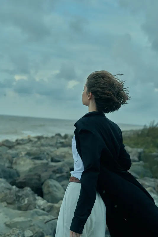Woman standing on rocky coastline, wind blowing her hair, back turned, wearing a dress and dark jacket, contemplative mood as sea and sky meet at the horizon