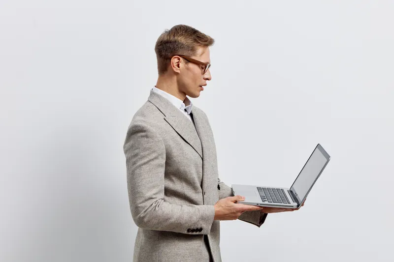 Professional young man in a gray suit and glasses holds a laptop computer, standing sideways against a plain light background, focused on work. Studio portrait of business professional, technology, an