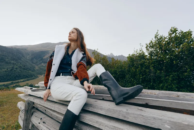 Woman in nature sits on wooden fence overlooking mountains, outdoor fashion with boots and warm jacket, relaxed pose in meadow landscape for travel and lifestyle.