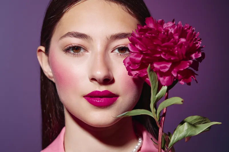 Close-up portrait of woman with glossy pink lips and smooth skin holding bright pink flower near her face against purple backdrop. Fashion concept in beauty creative style.