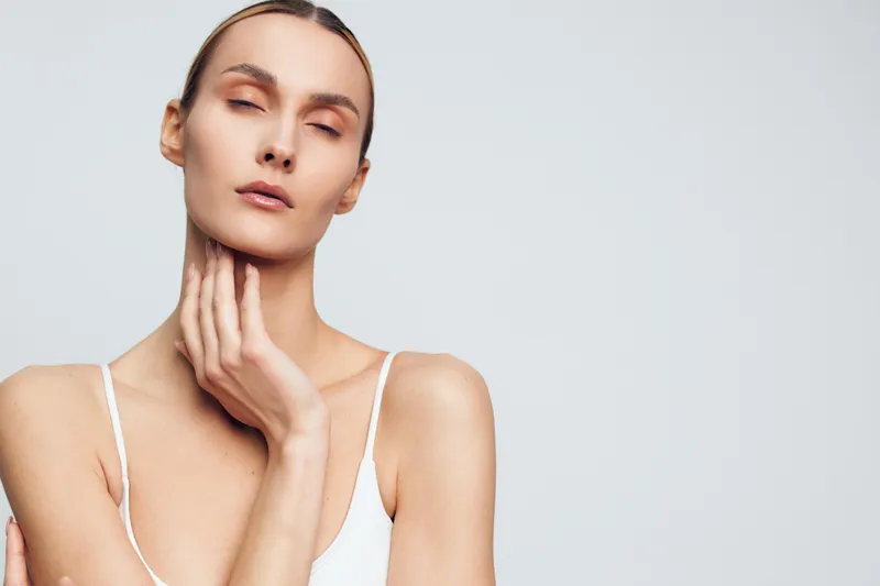 Portrait of a calm woman with closed eyes touching her neck, wearing a white tank top on a gray background, concept of beauty, skincare, wellness, and relaxation.