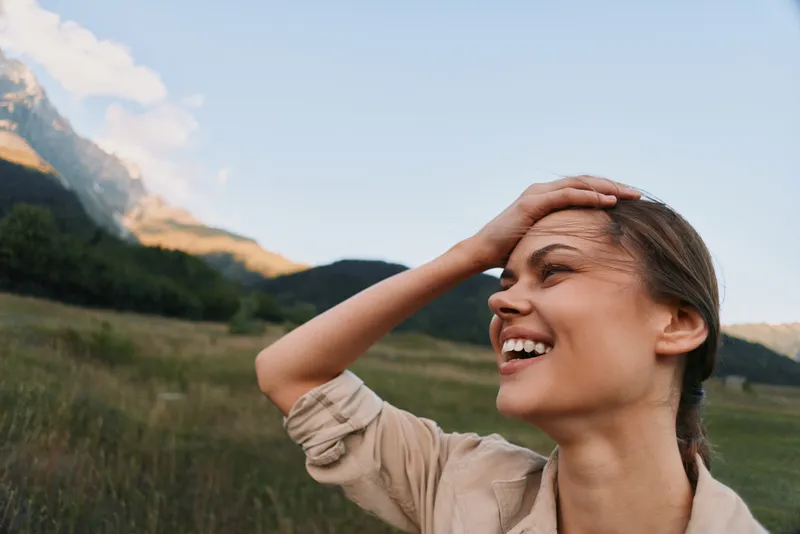Joyful woman enjoys outdoor lifestyle in a sunny field with distant mountains, smiling and carefree, capturing a candid moment of happiness and adventure.