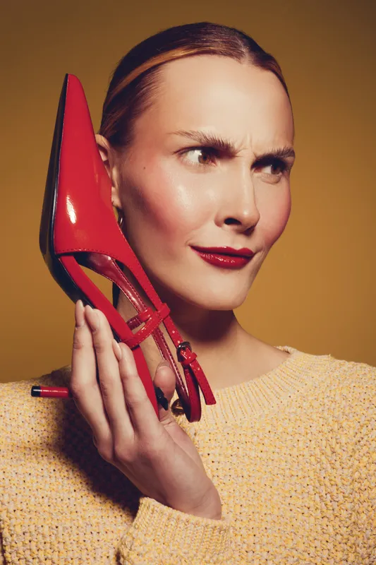 Woman in yellow sweater holds red high heel shoe near face with curious expression. Studio portrait on warm brown background. Concept for fashion, style, and footwear.