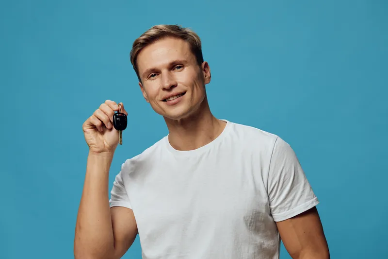 Smiling young man holding car key in hand, wearing casual white t-shirt, looking at camera, isolated on bright blue background. People lifestyle concept, automobile, transportation, happiness, fashion