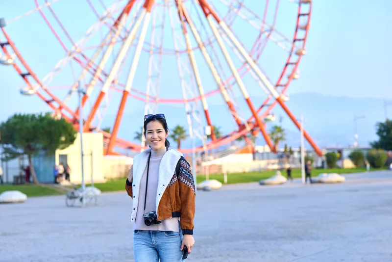 Travel photographer on a sunny day, smiling traveler with camera near a towering ferris wheel in a seaside park, casual fashion and candid outdoor photography mood