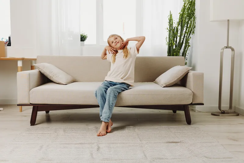 Relaxed girl in casual clothes sits barefoot on beige sofa stretching arms in bright living room with plants and natural light. Concept of comfort, home, and leisure time.