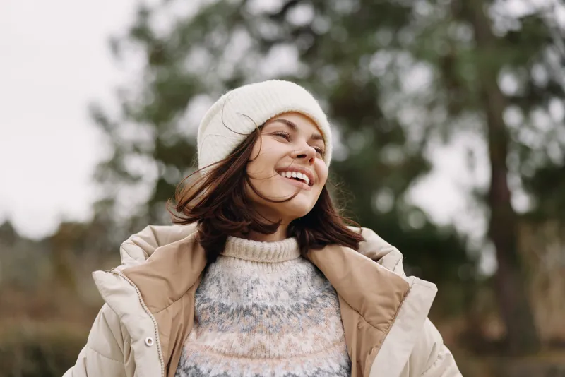 Outdoor woman smiling in a winter setting, wearing a beanie and a warm coat, a joyful portrait with soft natural light and blurred trees in the background