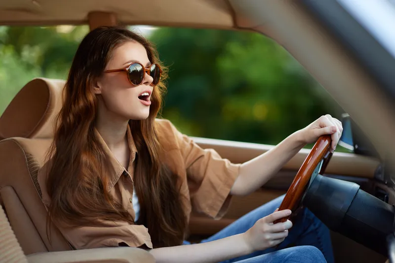 Young woman driving a car, singing joyfully while wearing sunglasses, showcasing vibrant outdoor energy and a sense of freedom on a sunny day.
