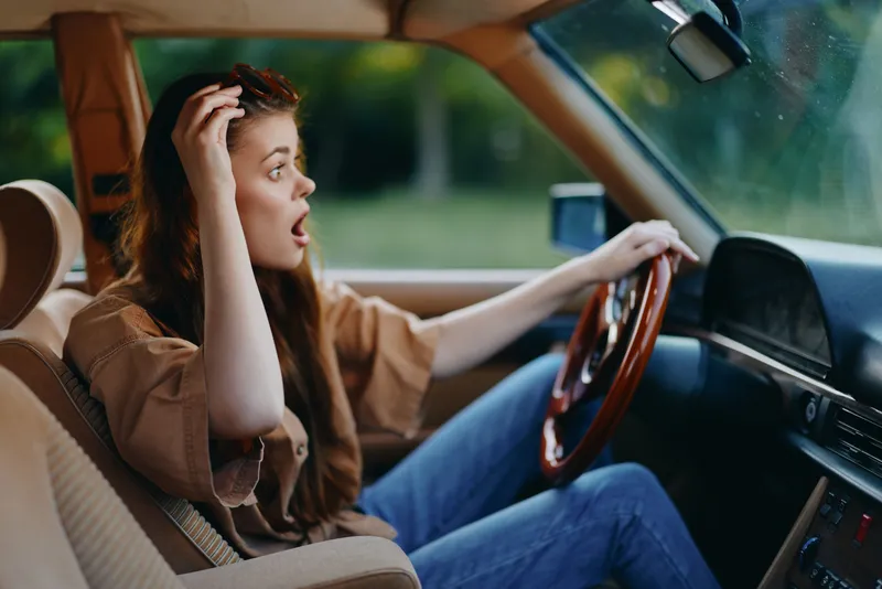 Surprised young woman driving a vintage car, wearing casual clothes, with a shocked expression against a natural green background.