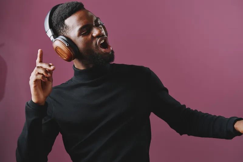 Young Black man enjoying music with headphones, expressive and joyful, in a stylish black turtleneck against a vibrant purple background