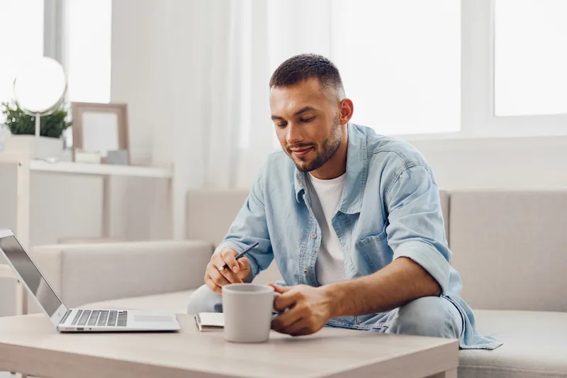 Stylish man enjoying a coffee while working on his laptop in a bright, modern living space. This scene captures the essence of remote work in a cozy environment.