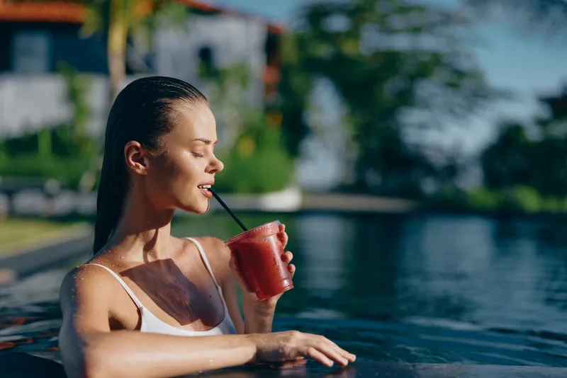 Young woman in white swimsuit relaxing in pool and drinking smoothie from plastic cup with straw. Sunny outdoor scene with blurred background of greenery and water.