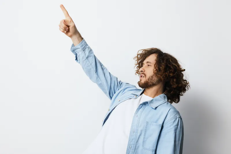 Young man in denim jacket pointing upwards with enthusiasm, showcasing emotions of joy and positivity in a minimalist white background