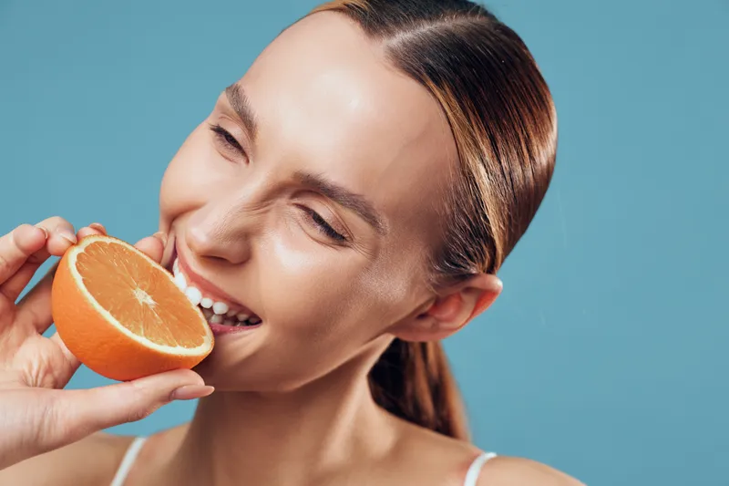 A smiling woman bites into a halved orange, fresh and bright against a blue background, conveying healthy eating and citrus flavor for wellness and vitality.