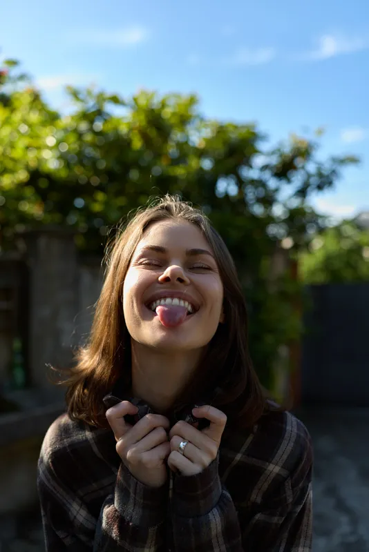 A joyful woman in casual plaid shirt outdoors, close up, playful with tongue out. Bright daylight and green foliage frame a carefree lifestyle moment