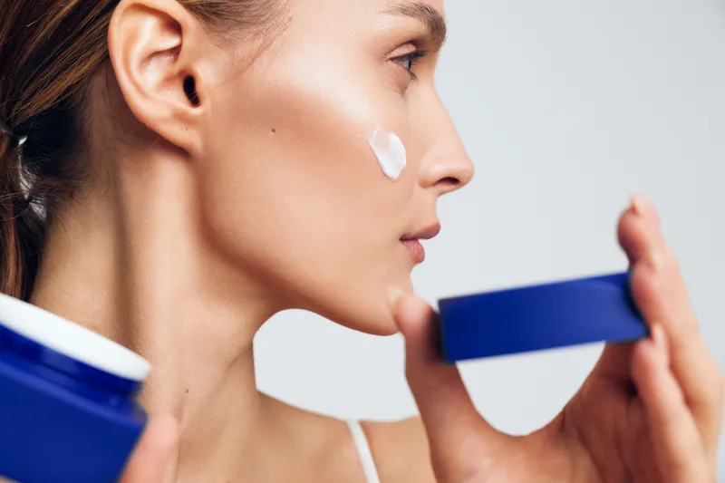 Young woman applies face cream on cheek holds blue jar skincare beauty routine natural skin care isolated white background closeup profile side view.