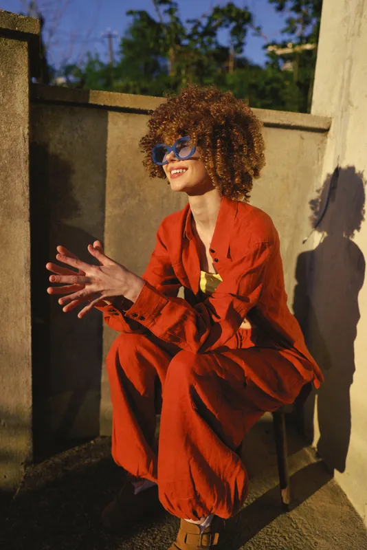 Vibrant woman lifestyle portrait outdoors on a sunny terrace, wearing an orange suit and sunglasses, smiling, relaxed pose, stylish and confident moment