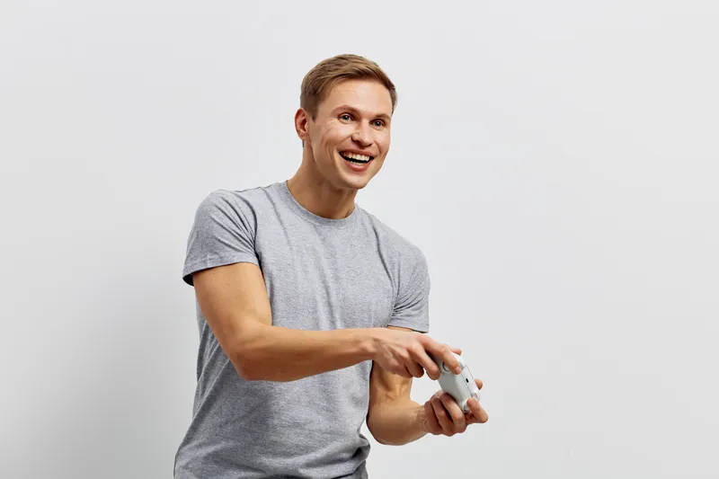 Smiling man in casual gray t-shirt holding game controller, expressing joy and excitement while playing video games, isolated on white background. Youth lifestyle leisure entertainment concept.