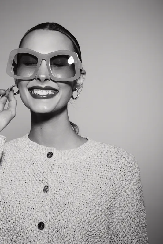 Smiling white woman with pale skin wearing oversized sunglasses and knitted sweater in studio black and white portrait.