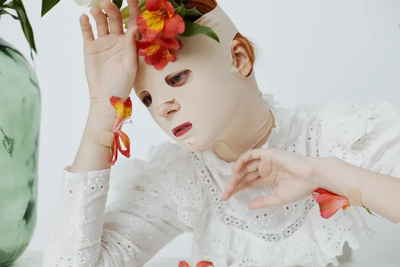 A woman wearing a floral face mask, expressing emotions of melancholy while surrounded by colorful flowers in a bright, minimalist setting