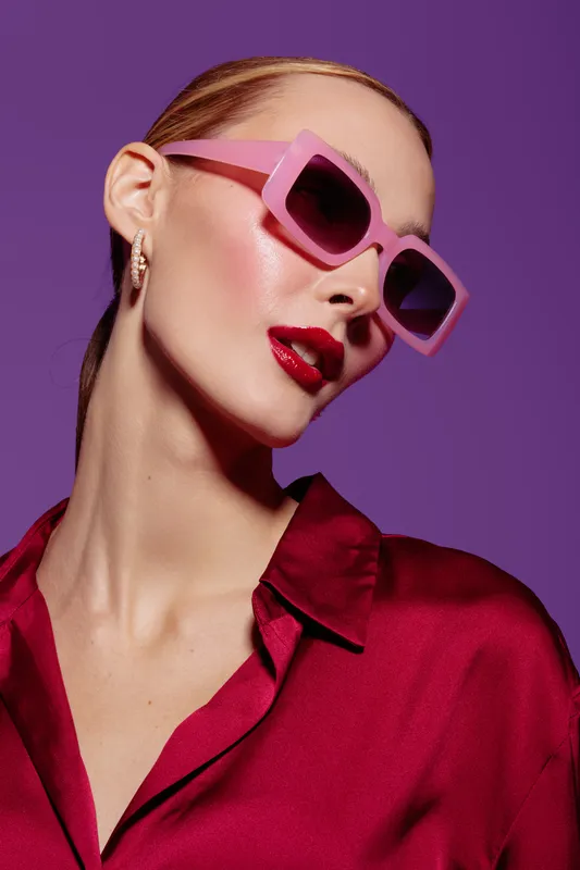 Stylish woman with pink sunglasses and red lipstick poses against purple background. She wears a red satin shirt and gold earrings. Concept of fashion, beauty, and glamour.