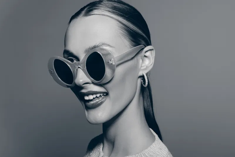 Smiling young white woman with light skin and slicked-back hair wearing large round sunglasses and earrings. Studio portrait with stylish fashion and confident mood.