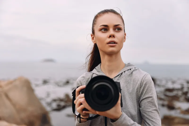 Young woman in a gray hoodie holding a camera at a coastal location, expressing curiosity and creativity while capturing nature s beauty