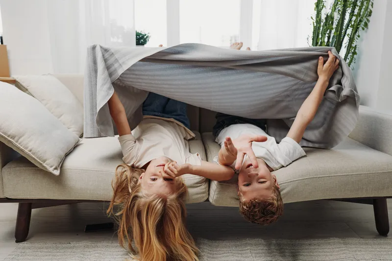 Happy mother and child lying upside down on a sofa covered with a blanket, smiling and playing indoors in a bright modern living room. Concept of family bonding and joy.