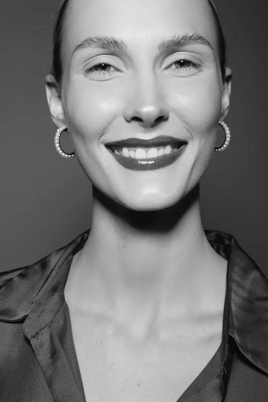 Closeup portrait of smiling white woman with light skin, wearing hoop earrings and dark blouse, isolated on black background.