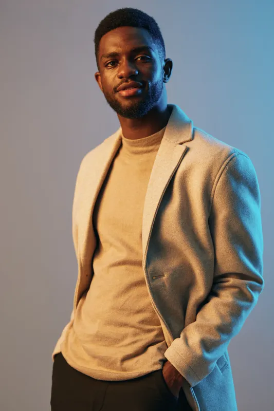 Stylish young Black man in a beige coat and sweater, evoking confidence and modern elegance against a soft blue background