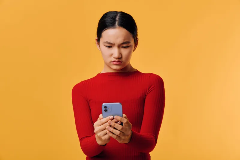 Young woman in red sweater holding smartphone with focused expression, using mobile device for texting or browsing on orange background in studio portrait.