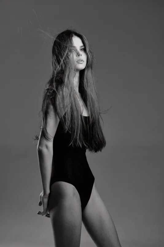 Black and white portrait of a woman with long hair wearing a black bodysuit, striking a thoughtful and elegant pose.