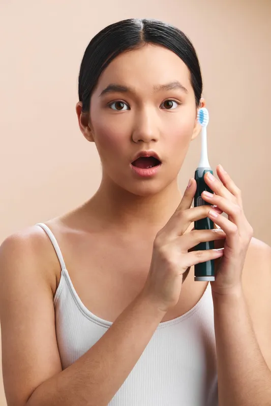 Asian woman shows surprise as she holds a toothbrush near her face in a bright bathroom, creating a playful candid moment that highlights daily dental care routine.