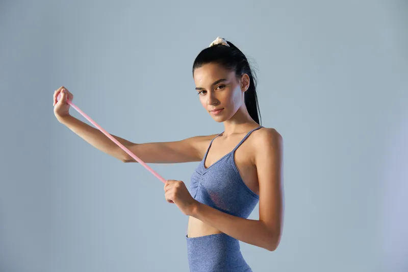 woman sport fitness stretching with resistance band, wearing blue workout outfit, focused young adult exercising indoors on neutral background