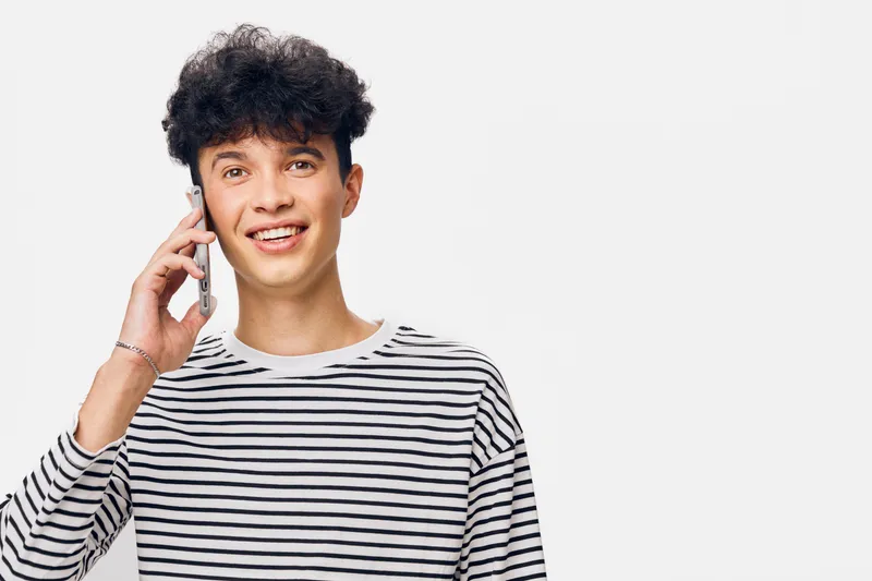 smiling young man talking on smartphone wearing casual striped shirt with curly hair on white background expressing happiness and friendliness.