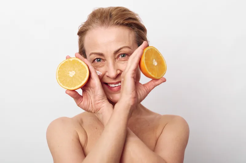 Smiling mature woman holds two halves of fresh orange near face, showing healthy skin and natural beauty on white background. Concept for skincare, wellness, and fruit therapy.
