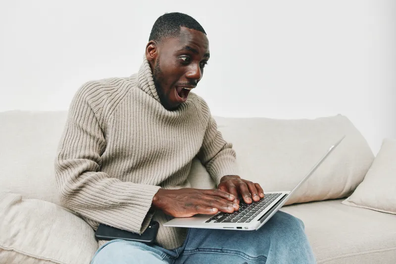 Excited young African American man in a cozy sweater using a laptop at home, expressing joy and surprise with a neutral background