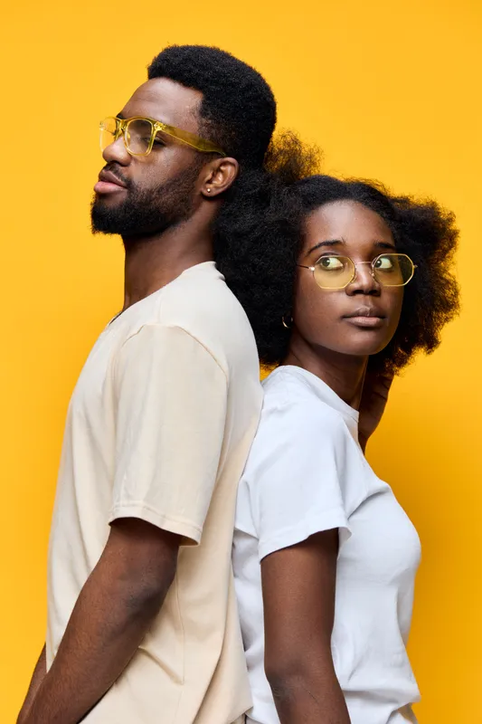 Young African couple wearing stylish glasses and casual clothing, posing back to back against a bright yellow background, showcasing modern and confident vibes
