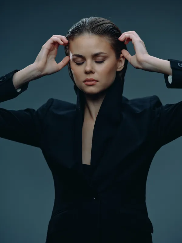 serious woman in black suit with closed eyes adjusting hair on gray background